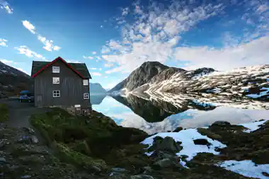 Dark wooden cabin beside still mountain lake with snow peaks
