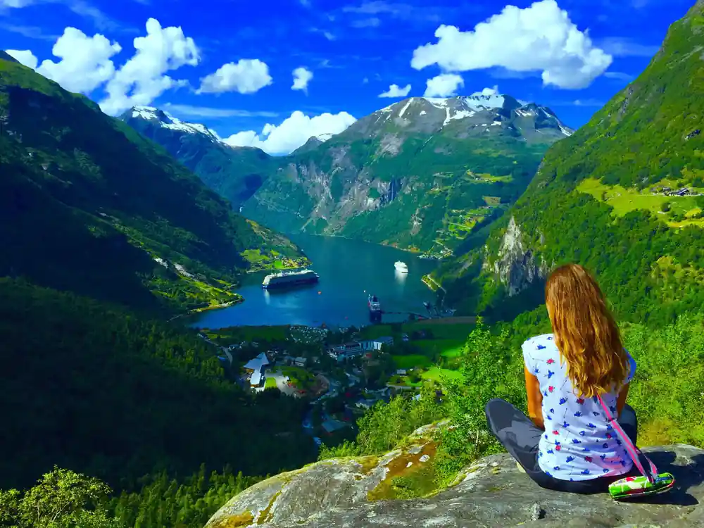 Woman sitting on rock overlooking cruise ship in deep blue fjord